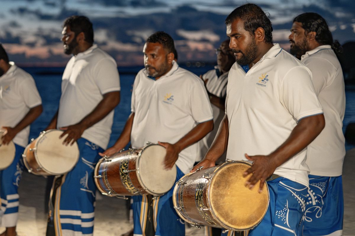 Group of Boduberu drummers dressed in traditional attire, performing near the beach at dusk. The musicians are holding large drums and wearing white shirts and blue sarongs with decorative patterns. The sky is darkening, creating a dramatic backdrop for the rhythmic and energetic performance.