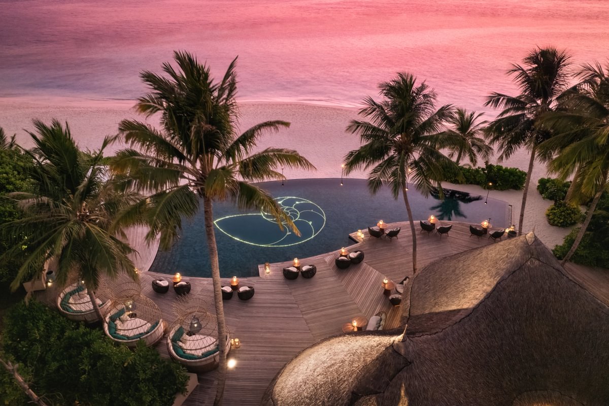 Aerial view of a poolside bar at sunset, surrounded by palm trees and overlooking the beach. The sky is painted in soft pink and purple hues, and there are intimate seating areas with tables and cozy chairs set up around the curved wooden deck. A glowing nautilus design is illuminated in the pool, adding to the tranquil and romantic atmosphere.